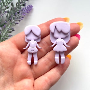 May include: Two purple resin doll charms, one larger than the other, with a simple design. The dolls have a skirt, legs, and hair.