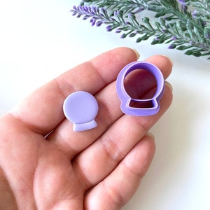 May include: Purple plastic cookie cutters shaped like a snow globe. The cutters are in the palm of a hand.