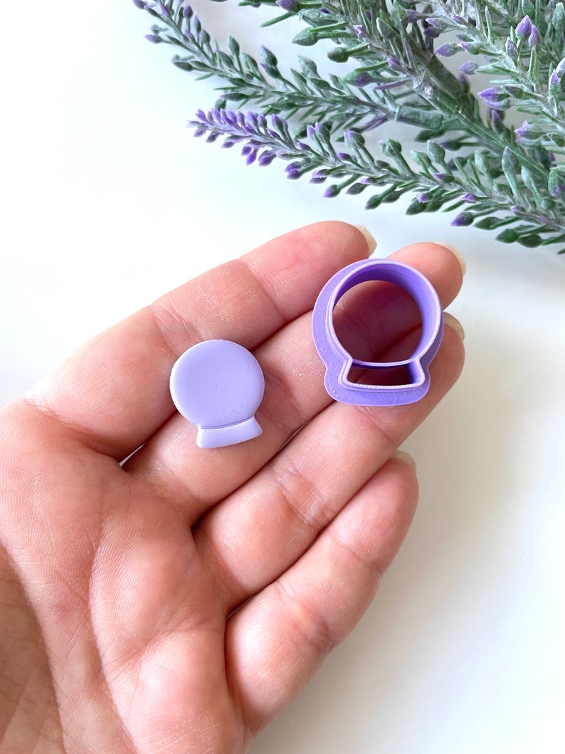 May include: Purple plastic cookie cutters shaped like a snow globe. The cutters are in the palm of a hand.
