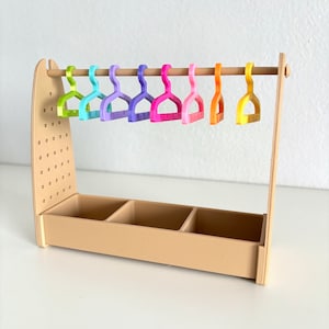 Può includere: Un appendiabiti in legno di dimensioni per case di bambole con una base marrone e un'asta in legno. L'appendiabiti ha tre scomparti nella base e otto grucce di plastica colorate appese all'asta.