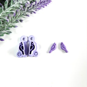 May include: Two purple bird-shaped polymer clay earrings with a heart detail and two smaller purple bird-shaped polymer clay earrings.
