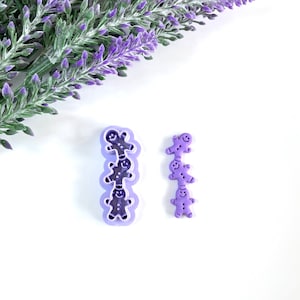 May include: Two purple plastic gingerbread man cookie cutters. One cutter is a single gingerbread man, the other is a stack of four gingerbread men.