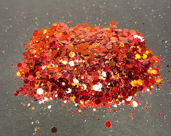 May include: A pile of red, orange, and gold glitter with a holographic finish. The glitter is scattered on a gray surface.