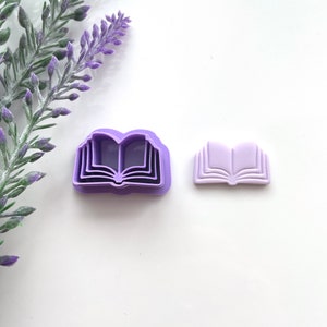 May include: Two purple plastic cookie cutters shaped like open books. The cutter on the left has multiple layers, while the cutter on the right is a single layer.