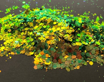 May include: A close-up of green and gold glitter, with a holographic finish. The glitter is scattered on a dark gray surface.
