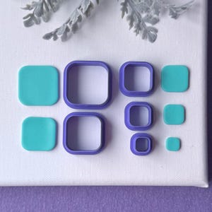 May include: A collection of square and rounded square cookie cutters in various sizes. The cutters are teal and purple, arranged on a white canvas with a purple background. A sprig of gray-green foliage is at the top.