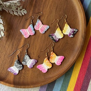 May include: Four pairs of butterfly-shaped dangle earrings featuring gradient colour schemes including pink, blue, yellow, purple, black, and orange. Each earring has a white line-art floral pattern and gold-toned metal hooks for attachment.