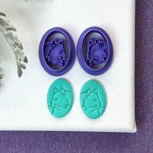 May include: Two purple plastic cat-shaped cookie cutters and two teal cat-shaped cookies.