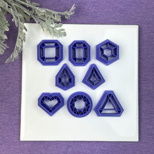 May include: A set of eight purple plastic cookie cutters shaped like different gemstones, including a heart, a circle, a triangle, and a square.