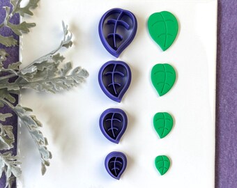 May include: Four purple leaf-shaped cookie cutters and four green leaf-shaped cookies are arranged on a white surface against a purple background. The cutters are in various sizes, with internal vein details. The green leaves are also in different sizes.