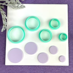 May include: A collection of circle-shaped polymer clay cutters and matching lilac-colored clay circles. The cutters are a vibrant teal color. The items are arranged on a white surface, with a purple background and a sprig of silver foliage.