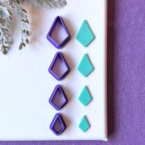 May include: A set of four purple diamond-shaped cutters arranged in a vertical column, accompanied by four corresponding teal diamond-shaped cutouts of decreasing sizes, displayed on a white surface next to a sprig of dusty miller foliage.