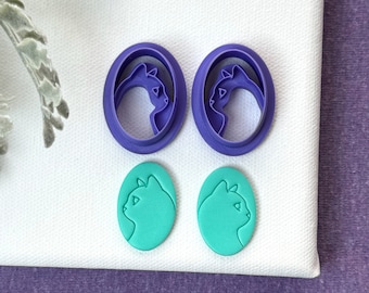 May include: Two pairs of cat-shaped earrings. One pair is purple and features a cat silhouette in a circle. The other pair is teal and features a cat silhouette in an oval.