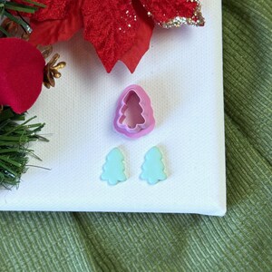 May include: A set of three pink and light blue plastic Christmas tree cookie cutters. The cutters are on a white surface.