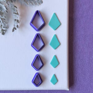May include: Four purple diamond-shaped polymer clay cutters and four teal diamond-shaped clay pieces are arranged on a white canvas. The cutters are hollow, while the clay pieces are solid. A sprig of gray foliage is in the upper left corner.