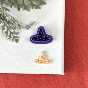 May include: Two witch hat cookie cutters, one purple and one brown, on a white surface.