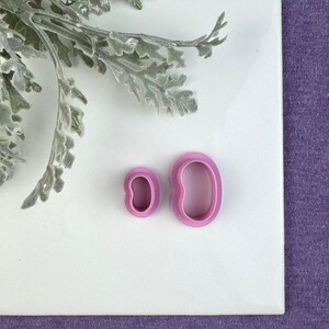 May include: Two pink plastic cookie cutters shaped like a bean, one larger than the other, on a white surface.