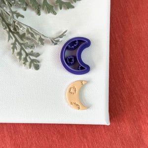 May include: Two crescent moon cookie cutters, one purple and one beige, on a white surface.