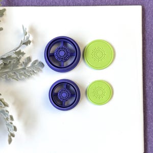 May include: Two purple and green cookie cutters. The purple cutters are circular with a wheel-like design. The green cutters are round with a starburst pattern. The cutters are on a white surface with a purple background.