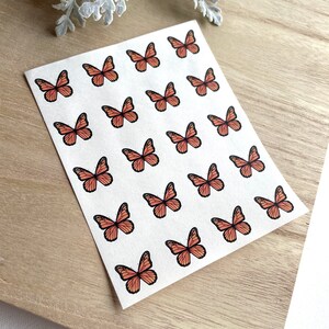 May include: A sheet of stickers featuring 20 orange butterfly designs. The butterflies are arranged in a grid pattern on a white background.