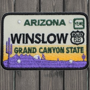 May include: Embroidered patch featuring the text "ARIZONA WINSLOW GRAND CANYON STATE" with a Route 66 logo. The design includes a purple desert landscape, a cactus, and the word "YEAR" in a green box.