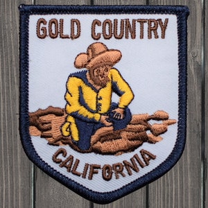 May include: A white embroidered patch with a blue border. It features a cartoon image of a prospector panning for gold in a riverbed. The text "GOLD COUNTRY CALIFORNIA" is embroidered in brown on the patch.
