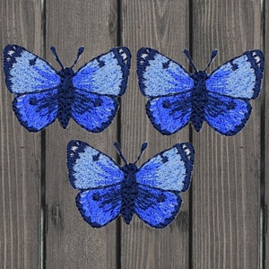 Puede incluir: Tres parches bordados de mariposas azules sobre un fondo de madera.
