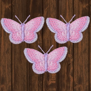 May include: Three pink and purple embroidered butterfly appliques. The butterflies have a light pink center and a darker pink border. The butterflies are on a brown wooden background.