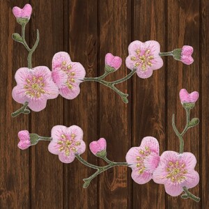 Puede incluir: Dos ramas de flores de cerezo bordadas en rosa con tallos y hojas verdes sobre un fondo de madera marrón.