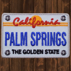 Può includere: Toppa ricamata bianca e grigia con un tramonto rosso e giallo, le parole "California" e "Palm Springs" in blu, e il testo "The Golden State" in nero.