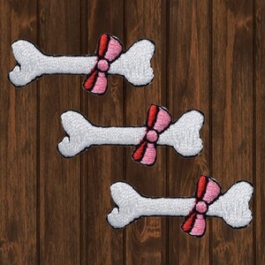May include: Three white bone-shaped patches with pink and red bows. The patches are embroidered and have a white background with pink and red stitching.