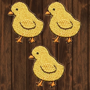 Puede incluir: Tres parches de pollito de fieltro amarillo con ojos y picos negros. Los pollitos están de pie sobre un fondo de madera.