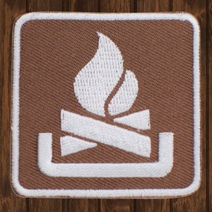 May include: A brown fabric patch with a white embroidered image of a campfire. The campfire is made up of three logs with a flame rising above them. The flame is stylized and has a teardrop shape.