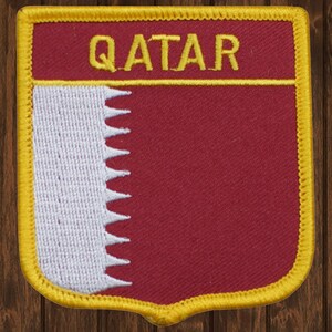 Puede incluir: Un parche bordado en color granate y blanco con un borde dorado. El parche presenta la palabra "QATAR" en letras doradas y la bandera de Qatar.