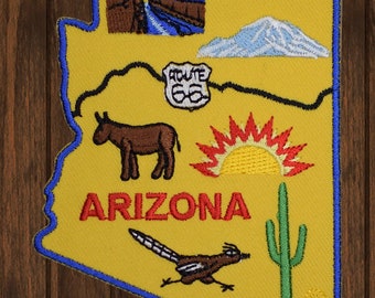 Puede incluir: Un parche de fieltro amarillo con forma de Arizona y un contorno azul. El parche presenta una salida del sol, un burro, un cactus, un correcaminos y un cartel de la Ruta 66. El texto "ARIZONA" está bordado en rojo.