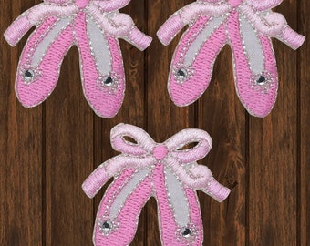 Op de afbeelding: Drie roze stoffen ballerina schoen applicaties met witte strikken en zilveren glitter accenten.