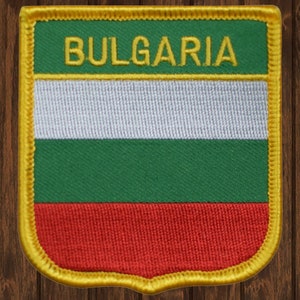 May include: Embroidered patch featuring the flag of Bulgaria. The patch has a shield shape with a gold border. The flag design includes horizontal stripes of white, green, and red, with the word "BULGARIA" in gold.