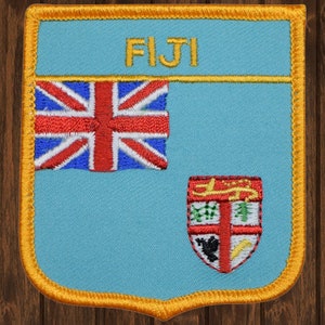 May include: A blue and gold embroidered patch with the word "FIJI" in yellow letters. The patch features the Fijian flag with the Union Jack in the upper left corner and the Fijian coat of arms in the lower right corner.