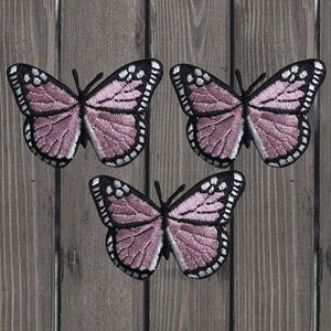May include: Three embroidered butterfly patches. Each butterfly has a mauve-colored wing with black and white detailing. The patches are arranged on a wooden background.