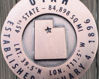 May include: A copper-colored round metal token with the word "UTAH" in black lettering at the top. The token also includes the text "45th STATE - 84,898 SQ MI", "LAT. 39.5°N LON. 111.5°W", "ESTABLISHED JANUARY 1896". The token features a black outline of the state of Utah with a star in the center.