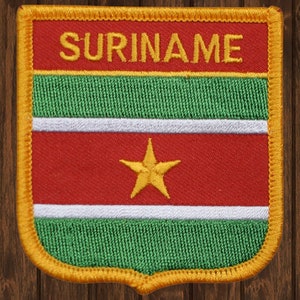 May include: A yellow embroidered patch with the word "SURINAME" in gold lettering and the Surinamese flag. The flag features a red field with a yellow five-pointed star in the center, bordered by two green stripes and a white stripe.