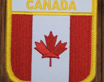 May include: A red and white embroidered patch with a maple leaf in the center and the word "CANADA" at the top. The patch has a yellow border.