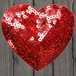 May include: A heart-shaped decoration covered in red sequins. The sequins are arranged to create a shimmering effect, with some areas appearing lighter due to the reflection of light. The background is a dark gray wooden surface.