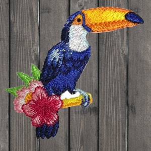 May include: Embroidered toucan patch. The patch depicts a toucan with a large orange beak, blue body, and white chest. It is perched on a yellow branch with pink and red flowers and green leaves. The patch is designed for clothing and accessories.