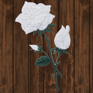 Op de afbeelding: Witte geborduurde roos met een knop en groene bladeren op een bruine houten achtergrond.