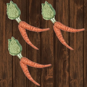 May include: Three felt carrots with green tops, arranged in a row on a brown wooden background.
