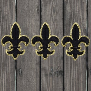 May include: Three black fleur-de-lis patches with gold embroidered trim. The patches are arranged in a row against a weathered wood background. Each patch features a detailed design with a classic fleur-de-lis shape.