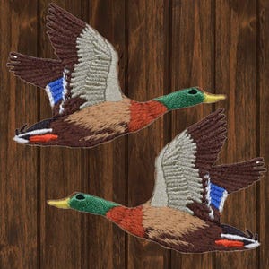 May include: Two embroidered patches of mallard ducks in flight. The ducks are brown, gray, green, and orange. The patches are on a wood background.