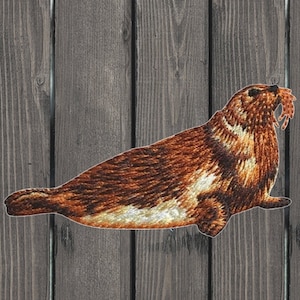 May include: Embroidered patch of a brown and white seal with its mouth open, as if it is eating something. The seal is lying on a wooden surface.