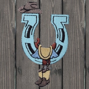 May include: A blue horseshoe with a cowboy boot and hat hanging from it. The horseshoe is embroidered with a brown and white pattern.